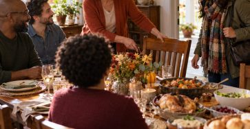 A warm diverse adult gathering at a modest autumn dinner table, an older woman pulling an extra chair toward a newly arriving adult neighbor, welcoming scene