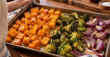 A metal sheet pan with separated groups of golden roasted butternut squash cubes, broccoli and red onion, autumn kitchen ingredients nearby