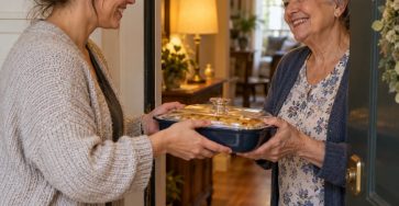 An adult woman and an older woman smiling at an apartment doorway as they exchange a covered casserole dish, warm hallway light, genuine neighborly kindness